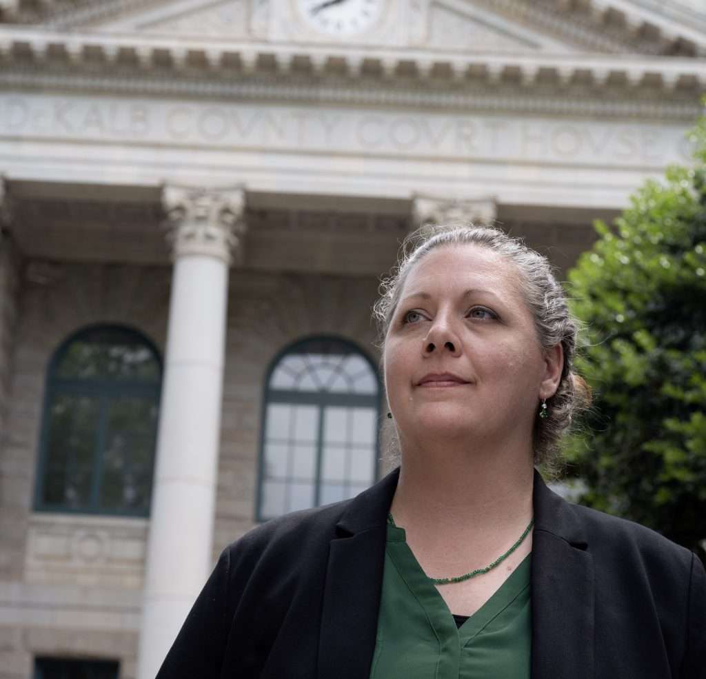 Kristen Howe is standing in front of an old courthouse building with large white columns and a clock at the top. She are wearing a black blazer over a green shirt and are looking determinedly into the distance. Trees frame the sides of the image.